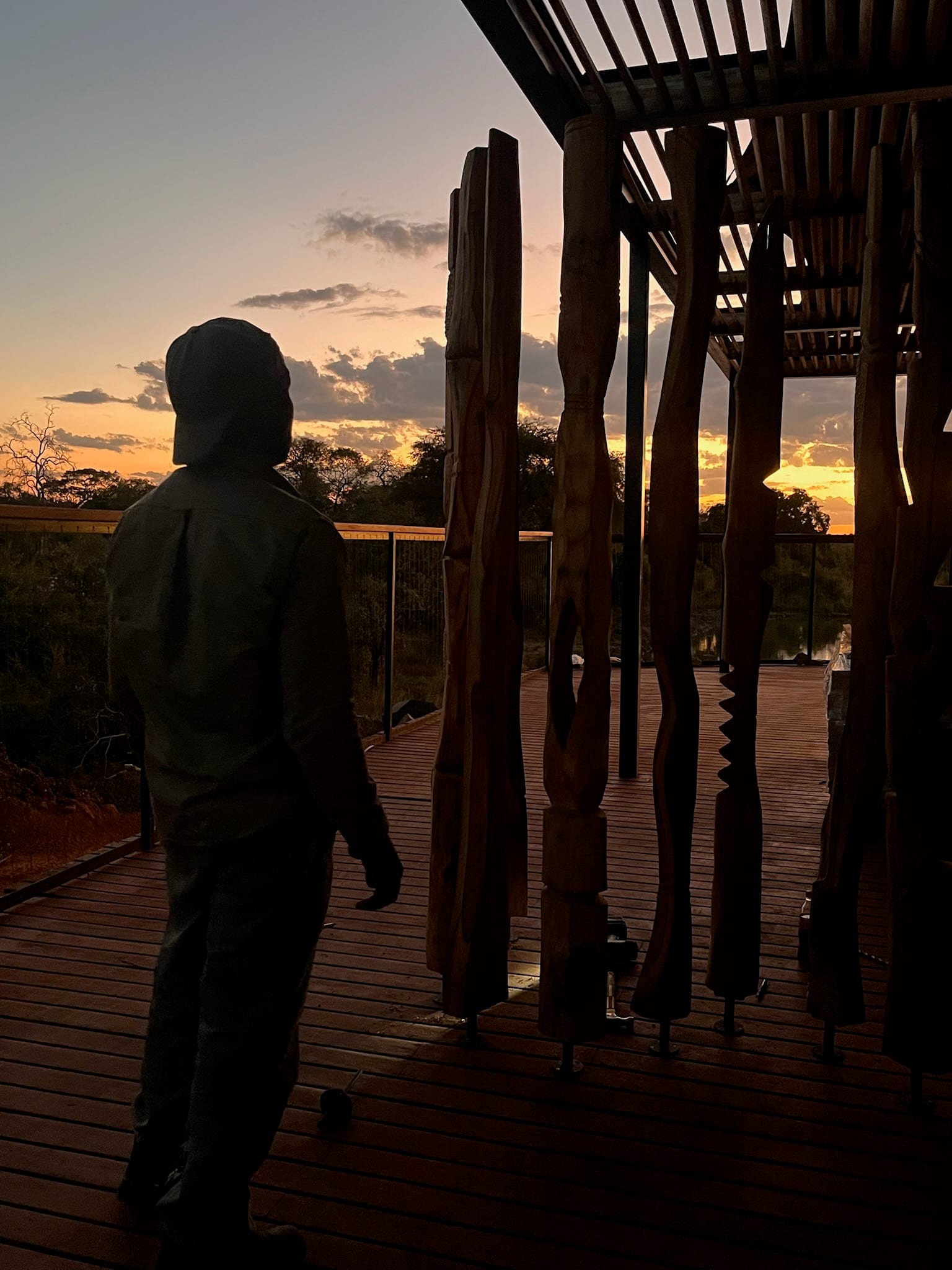 A timber deck under a thatched roof, looking onto acacia trees and open bush.
