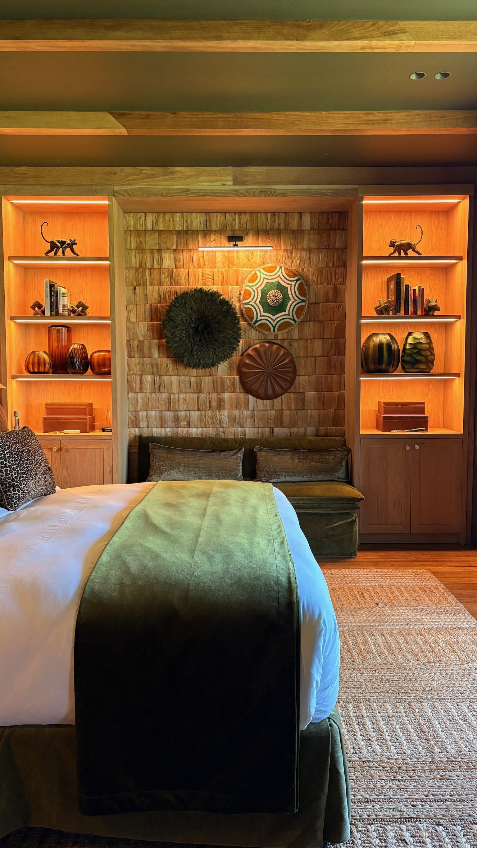 A bedroom with a green throw on the bed, timber shelves lit from within, and a woven wall.