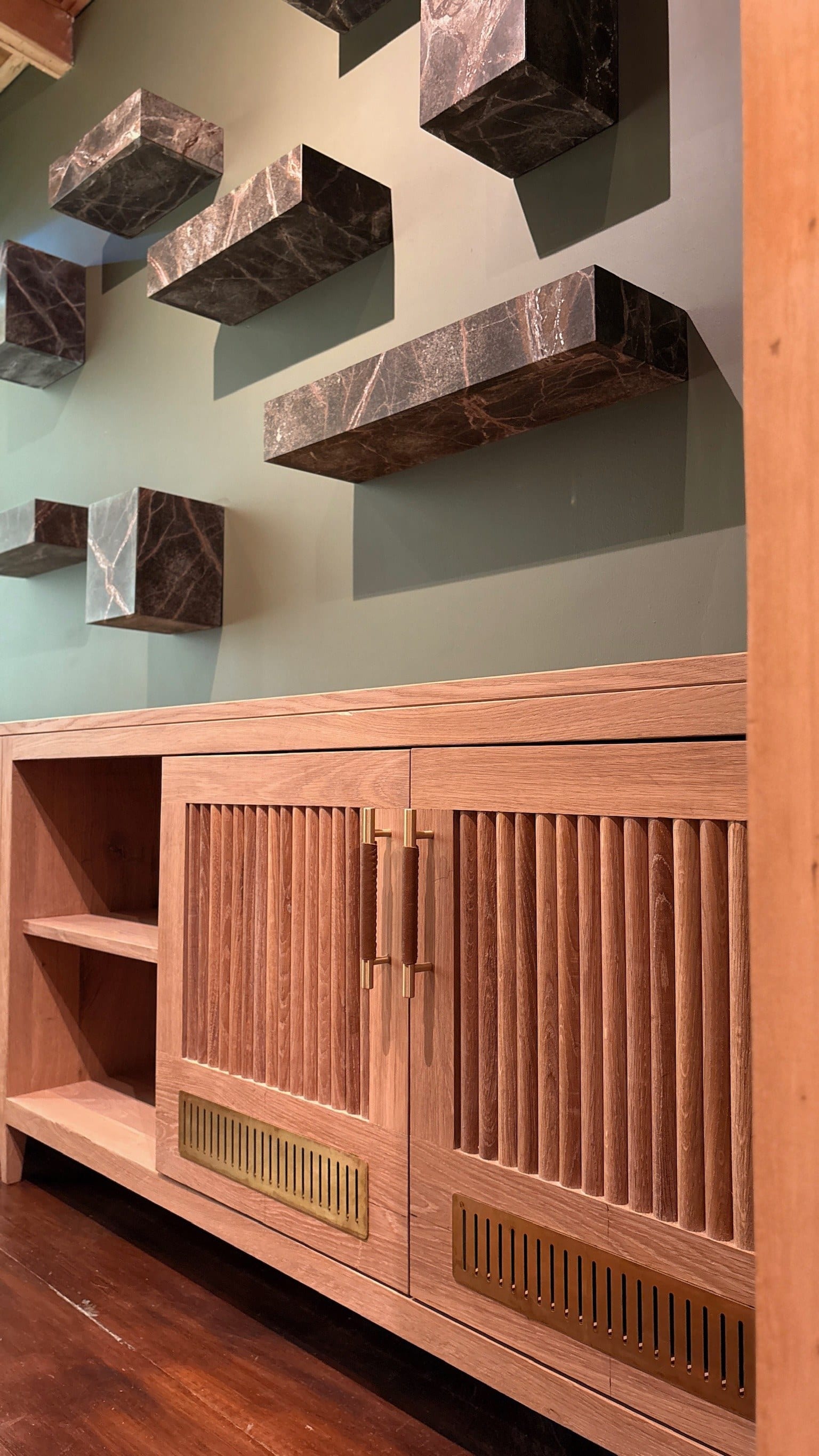 A timber sideboard with fluted doors, brass pulls and vents, under dark stone shelves on a sage wall.